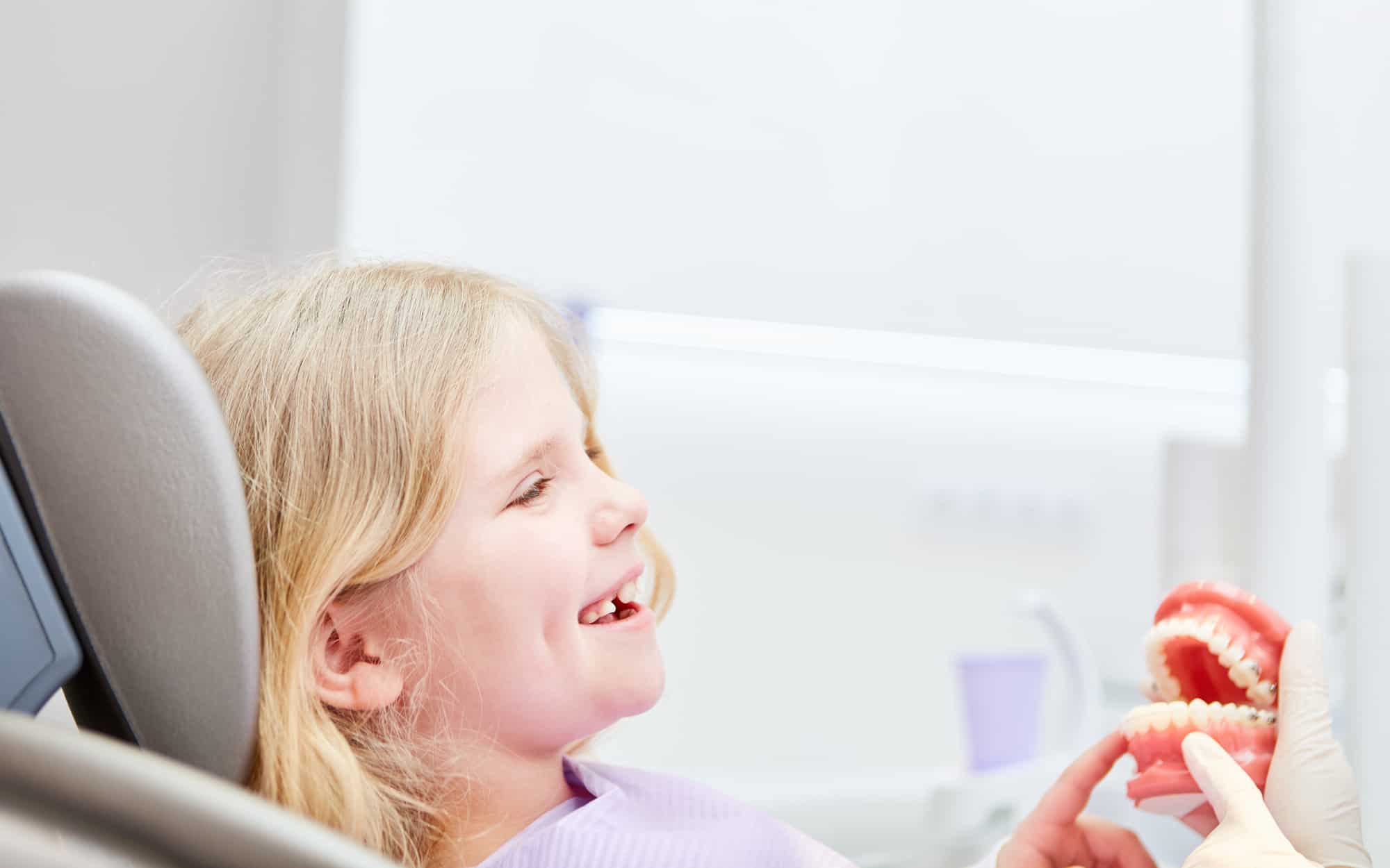Unser Ziel ist Ihre lebenslange Zahn- und Mundgesundheit - auch in der Kinderprophylaxe - Fröhliches Mädchen als Patient sitzt angstfrei beim Zahnarzt